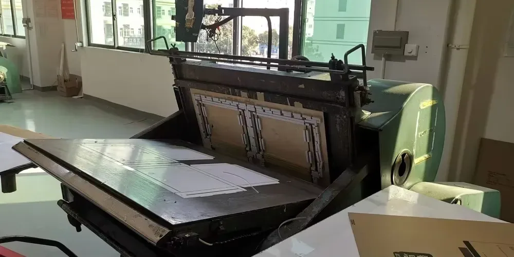 Cutting die locked in the platen press, shaped for a precise corrugated scratcher outline.